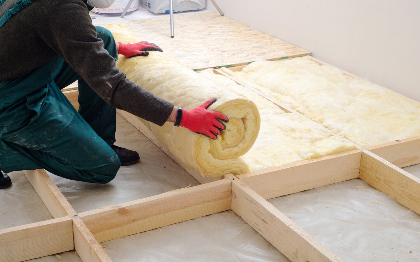 A person wearing long sleeves, overalls, and work gloves lays out fiberglass insulation under a floor.