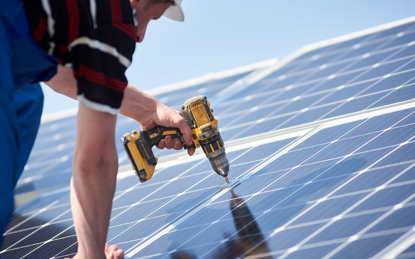 A white man in a striped shirt and blue pants uses a drill to install solar panels.