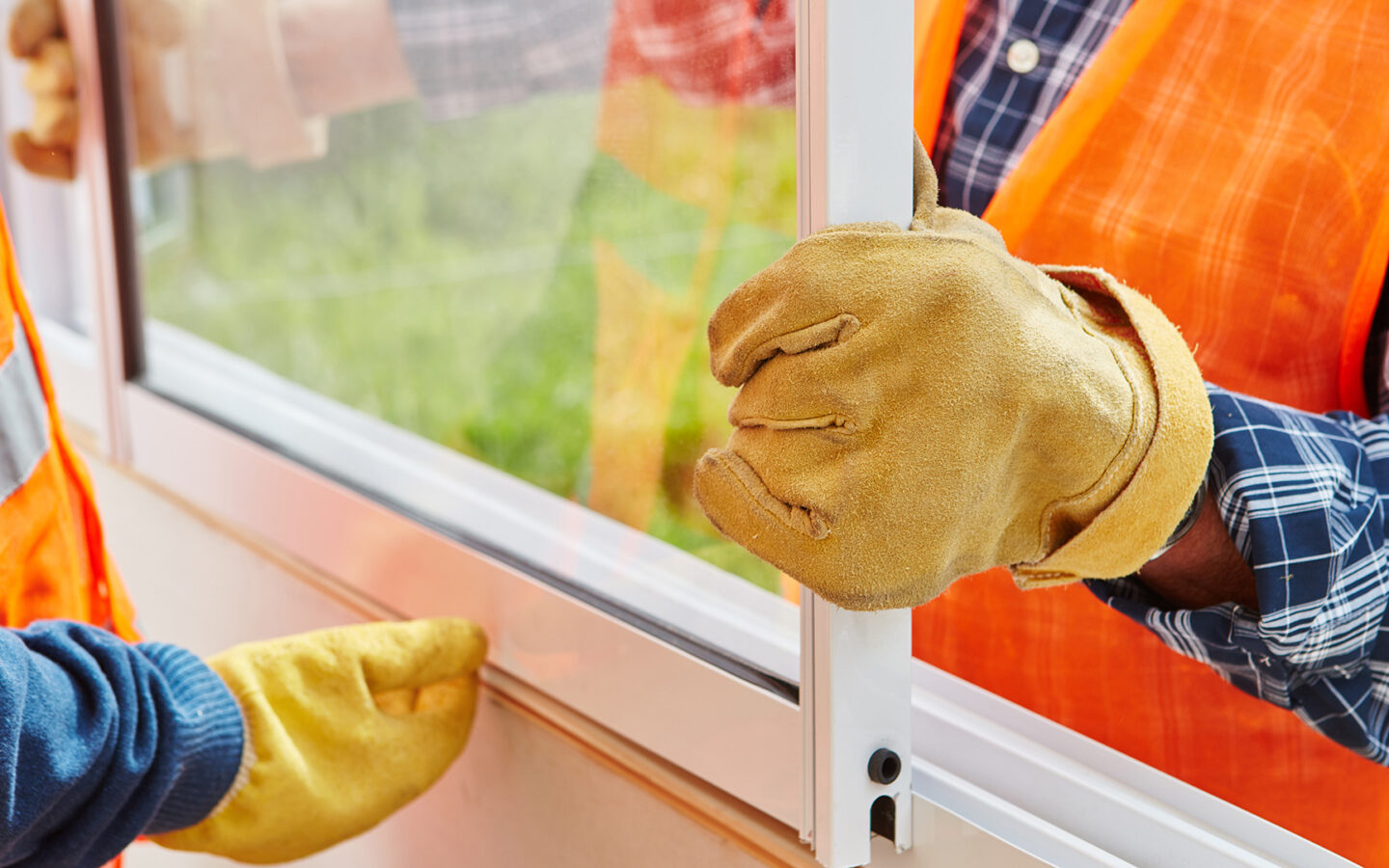 Two workers installing a new window in a home. The image is cropped to show torsos, hands, and arms as they work. They are wearing orange construction vests, work gloves, and long sleeve shirts.