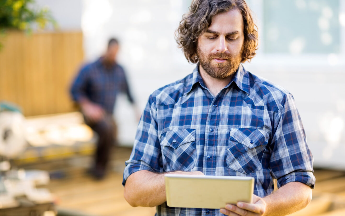 A white man with medium curly hair and a beard wearing a blue flannel shirt is working on a tablet. Blurred in the background is another man working on a construction project.